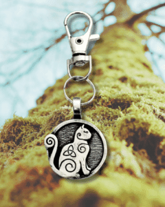 The Celtic Cat Bag Charm from the Celtic Spirals Collection features a silver keychain with a round cat and knot design, shown hanging before a mossy tree trunk with blurred branches in the background.