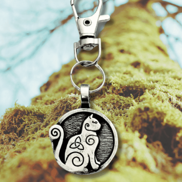 The Celtic Cat Bag Charm from the Celtic Spirals Collection features a silver keychain with a round cat and knot design, shown hanging before a mossy tree trunk with blurred branches in the background.