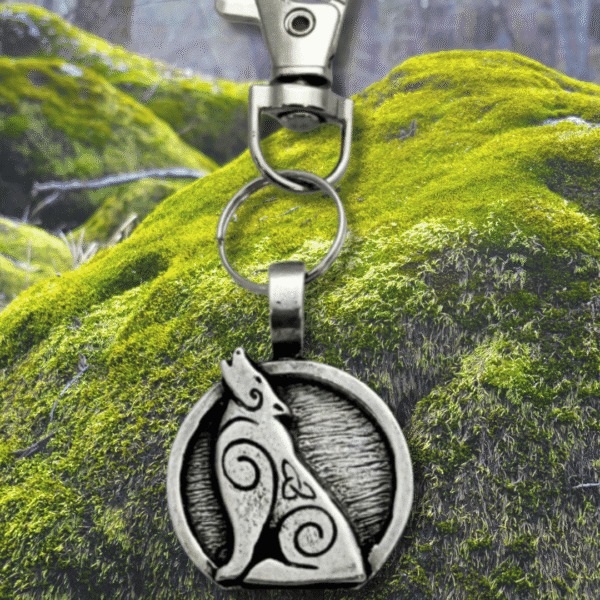 The Howling Wolf Bag Charm from the Celtic Spirals Collection features a silver keychain with a circular pendant of a howling wolf and Celtic swirls, perfect for accessorizing bags or keys against any forest-inspired backdrop.