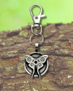 A silver keychain with a round pendant featuring an ornate butterfly design rests on rough tree bark, accompanied by the Luna Moth Bag Charm from the Celtic Spirals Collection, against a blurred green background.