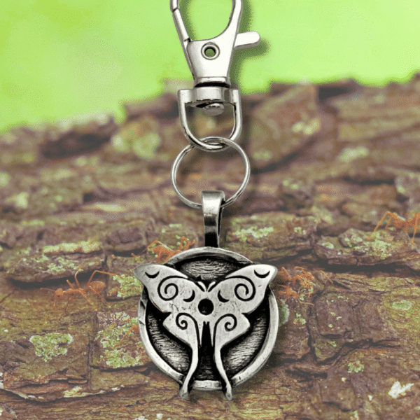 A silver keychain with a round pendant featuring an ornate butterfly design rests on rough tree bark, accompanied by the Luna Moth Bag Charm from the Celtic Spirals Collection, against a blurred green background.