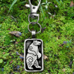 The Standing Bear Bag Charm from the Celtic Spirals Collection features an engraved bear with spiral designs, resting on green moss and leaves—perfect as a unique bag bauble.