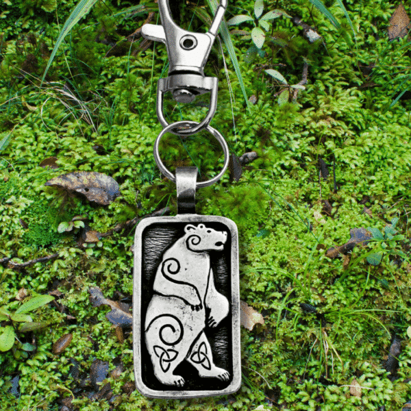 The Standing Bear Bag Charm from the Celtic Spirals Collection features an engraved bear with spiral designs, resting on green moss and leaves—perfect as a unique bag bauble.