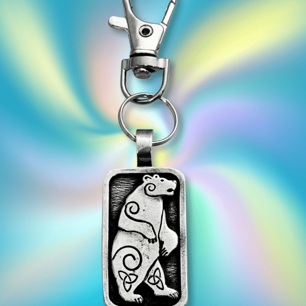 The Standing Bear Bag Charm from the Celtic Spirals Collection features a silver rectangular pendant with an engraved bear, swirling and Celtic knot designs, and a vibrant multicolored background—a unique adornment for bags or keys.