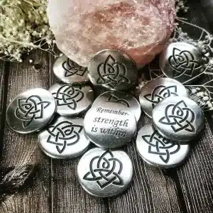 A 36 piece pack of Brigid's Heart Blessing Charms, featuring silver stones with triquetra symbols and one engraved “Remember: strength is within,” displayed on a wooden surface beside a pink crystal.