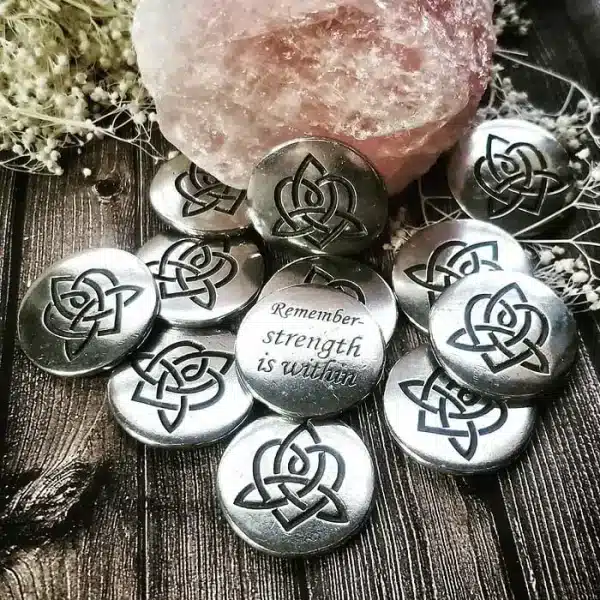 A 36 piece pack of Brigid's Heart Blessing Charms, featuring silver stones with triquetra symbols and one engraved “Remember: strength is within,” displayed on a wooden surface beside a pink crystal.