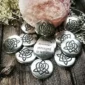 A 36 piece pack of Brigid's Heart Blessing Charms, featuring silver stones with triquetra symbols and one engraved “Remember: strength is within,” displayed on a wooden surface beside a pink crystal.