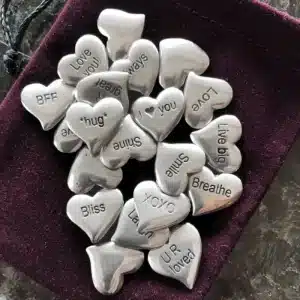 A bag of 100 Tiny Hearts Pocket Charms—small silver heart shaped stones with messages like “Love you,” “BFF,” “Smile,” “Bliss,” and “U R loved”—is displayed on a soft burgundy pouch.