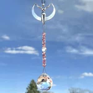The Crystal Serendipity Suncatcher features a silver angel with outstretched wings holding a crescent moon, and below, pink beads and a faceted crystal ball dangle against a blue sky dotted with clouds.