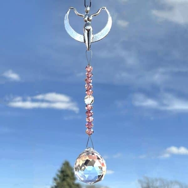 The Crystal Serendipity Suncatcher features a silver angel with outstretched wings holding a crescent moon, and below, pink beads and a faceted crystal ball dangle against a blue sky dotted with clouds.