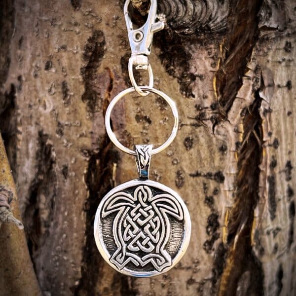 A Celtic Turtle Bag Charm with a round pendant showcasing a Celtic knot turtle design hangs from a silver keychain on a tree trunk.