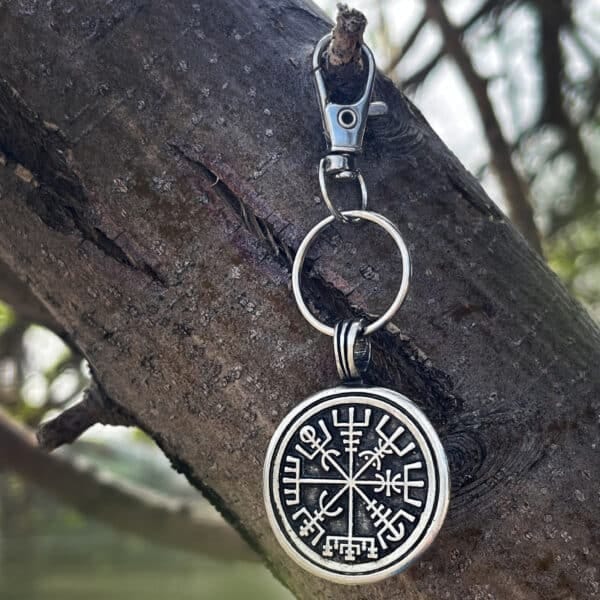 The Wayfinder Bag Charm, a round metal keychain with a Norse Vegvisir symbol in black and silver and intricate rune-like details, hangs from a tree branch as sunlight filters through blurred greenery.