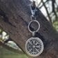 The Wayfinder Bag Charm, a round metal keychain with a Norse Vegvisir symbol in black and silver and intricate rune-like details, hangs from a tree branch as sunlight filters through blurred greenery.