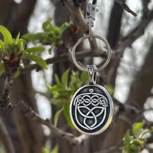 The Celtic Acorn Bag Charm, featuring a silver Celtic knot design, hangs from a budding tree branch with fresh green leaves, with more branches softly blurred in the background.