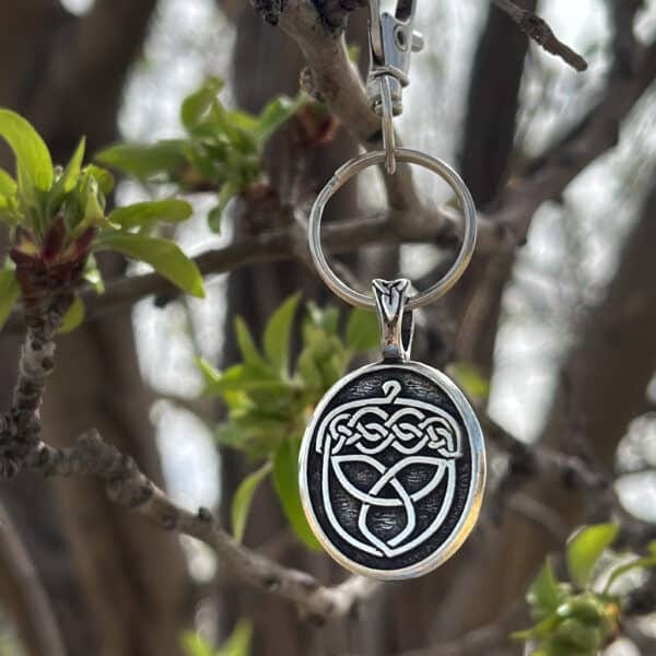 The Celtic Acorn Bag Charm, featuring a silver Celtic knot design, hangs from a budding tree branch with fresh green leaves, with more branches softly blurred in the background.