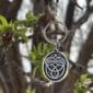 The Celtic Acorn Bag Charm, featuring a silver Celtic knot design, hangs from a budding tree branch with fresh green leaves, with more branches softly blurred in the background.