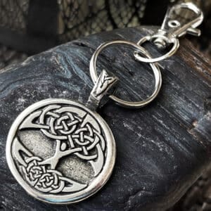 A Celtic Tree of Life Bag Charm with a circular pendant and silver keyring rests on a textured dark rock surface. The lobster clasp is attached to the pendant’s loop.