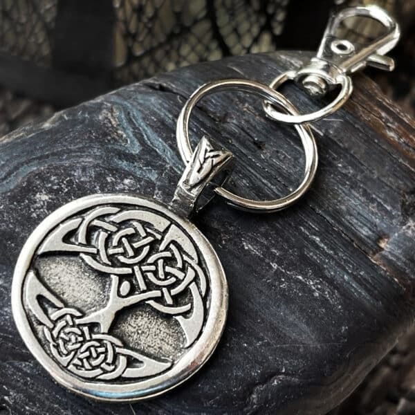 A Celtic Tree of Life Bag Charm with a circular pendant and silver keyring rests on a textured dark rock surface. The lobster clasp is attached to the pendant’s loop.