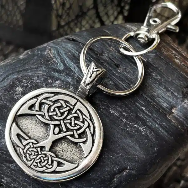 A Celtic Tree of Life Bag Charm with a circular pendant and silver keyring rests on a textured dark rock surface. The lobster clasp is attached to the pendantโs loop.