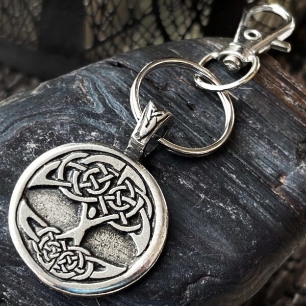 A Celtic Tree of Life Bag Charm with a circular pendant and silver keyring rests on a textured dark rock surface. The lobster clasp is attached to the pendantโs loop.