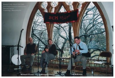 Three musicians perform on a stage in front of large arched windows with trees outside. Two sit with microphones; one plays uilleann pipes. A banner above reads "BCM Fest." Instruments and chairs are on stage.